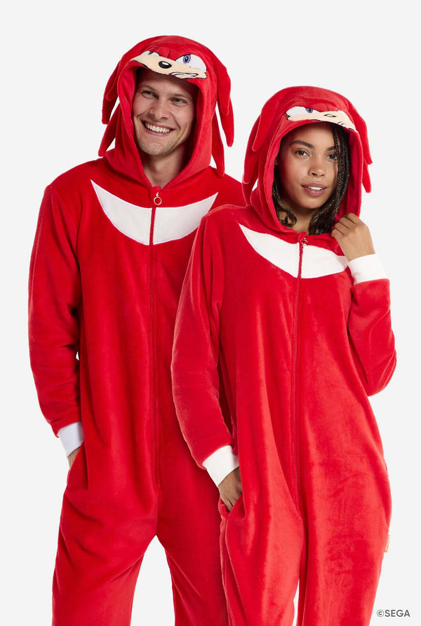 A man and woman are wearing a bright red Knuckles the Echidna-themed adult onesie from OppoSuits. The fun and cozy onesie features a zip-up front and an attached hood designed to resemble Knuckles' face, complete with expressive eyes and floppy dreadlocks. The suit is primarily red, with a white crescent pattern on the chest, reflecting Knuckles' iconic look from the Sonic the Hedgehog video game series. 