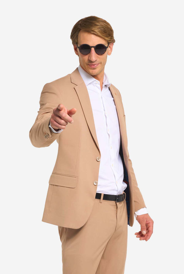 Image with light grey background and man wearing sunglasses, Dark Sand men’s suit and white shirt. Suits consists of blazer and pants. Pointing to the camera.