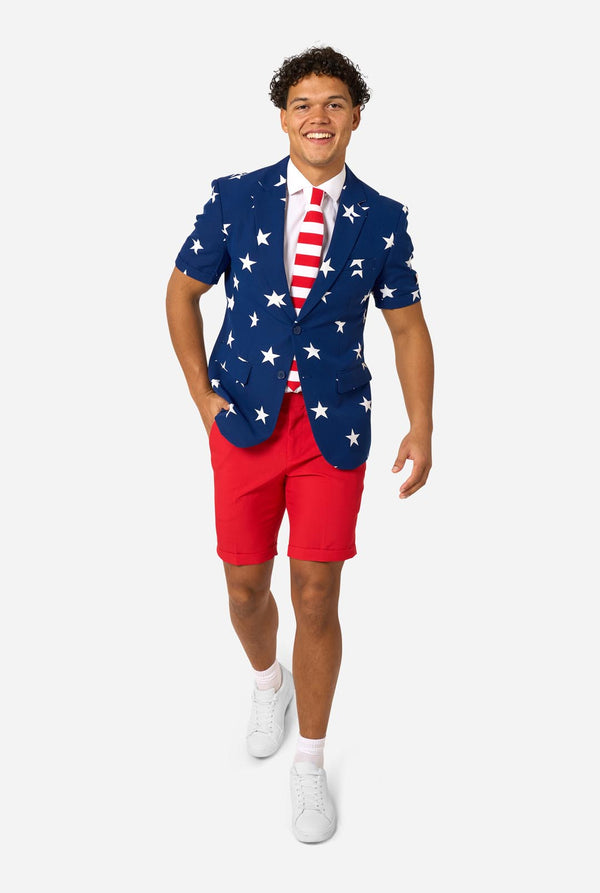 Person wearing a blue blazer with white star patterns, white shirt, and red shorts on a white background. Full body image