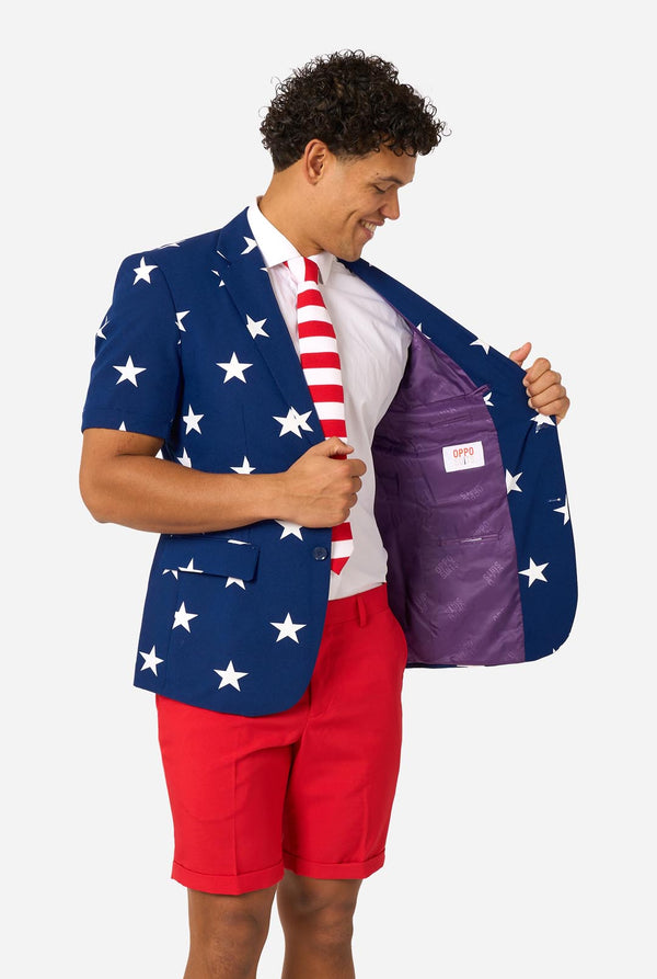 Person wearing a blue blazer with white star patterns, white shirt, and red shorts on a white background. Holding one side of the jacket open.