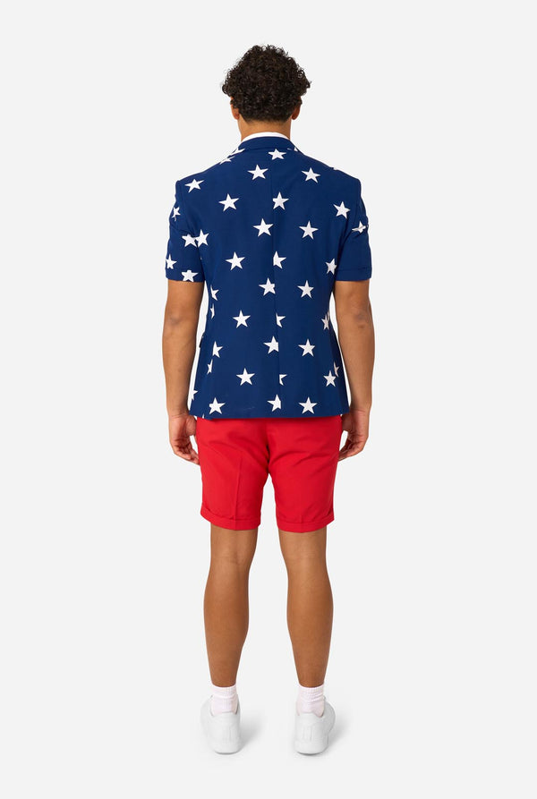 Person wearing a blue blazer with white star patterns, white shirt, and red shorts on a white background. View from the back.