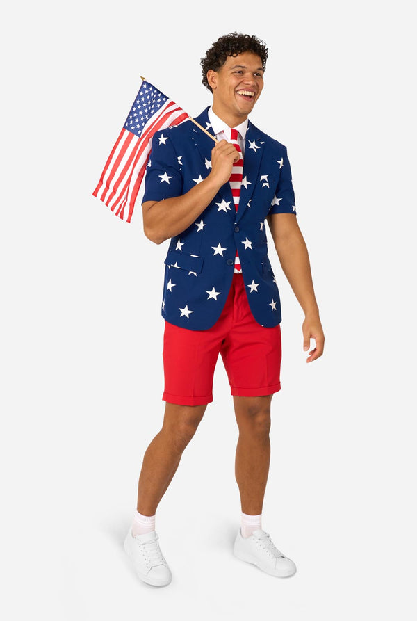 Person wearing a blue blazer with white star patterns, white shirt, and red shorts on a white background and carying a small usa flag.