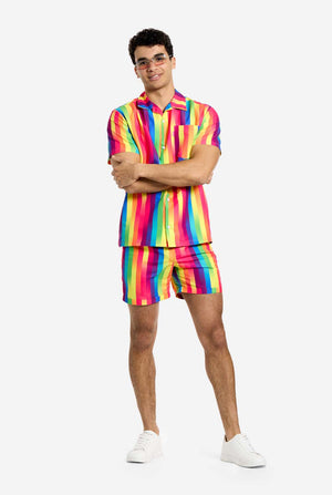 A man is wearing a vertically striped rainbow-colored summer set, consisting of shirt and shorts. He is smiling in the camera and holding his arms crossed. Also wearing sunglasses.