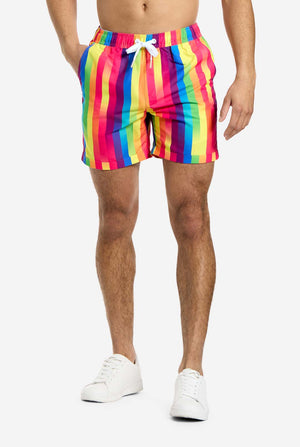 A man is wearing vertically striped rainbow-colored shorts. He is holding one hand in his pocket. Only showing the bottom part of his body.