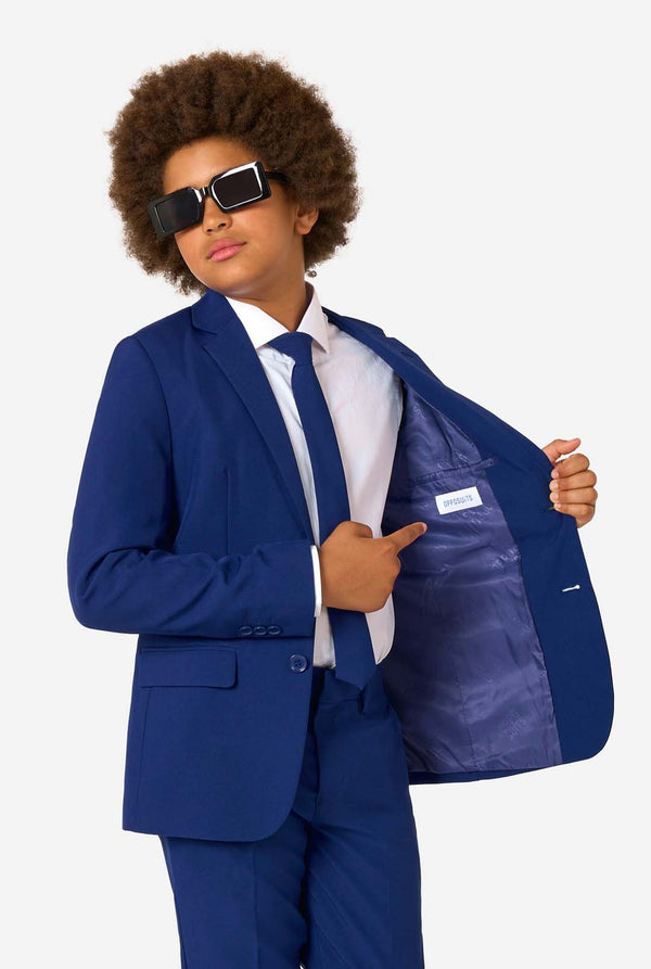 Kid wearing a blue suit with sunglasses, pointing to the jacket pocket on a white background
