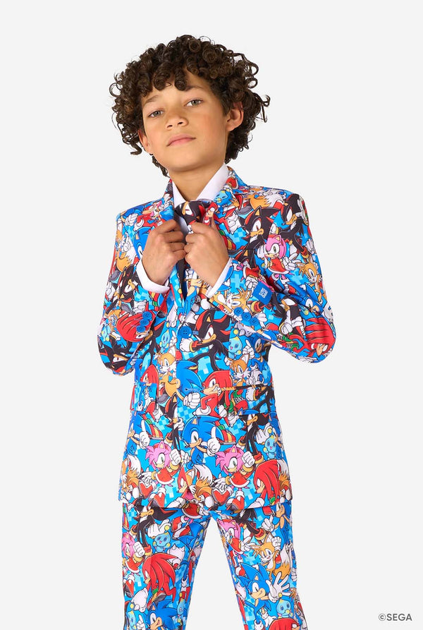 Young boy wearing a vibrant Sonic teen suit featuring colorful prints of Sonic the Hedgehog characters including Sonic, Knuckles, Tails, and Amy, paired with a matching tie and white shirt. Looking in the camera.