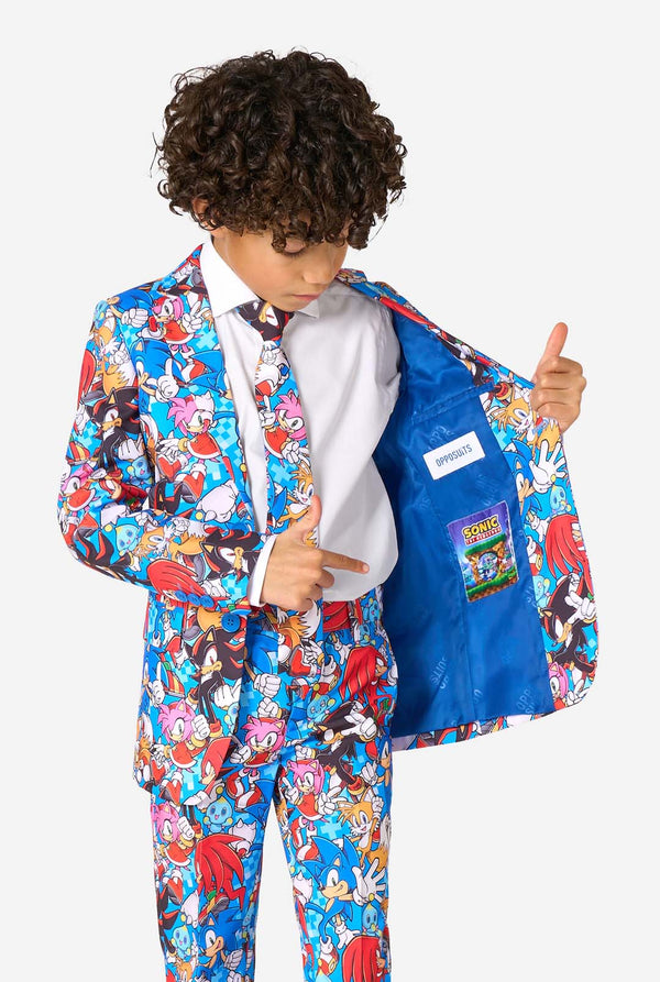 Young boy wearing a vibrant Sonic teen suit featuring colorful prints of Sonic the Hedgehog characters including Sonic, Knuckles, Tails, and Amy, paired with a matching tie and white shirt. Holding one side of his jacket open.