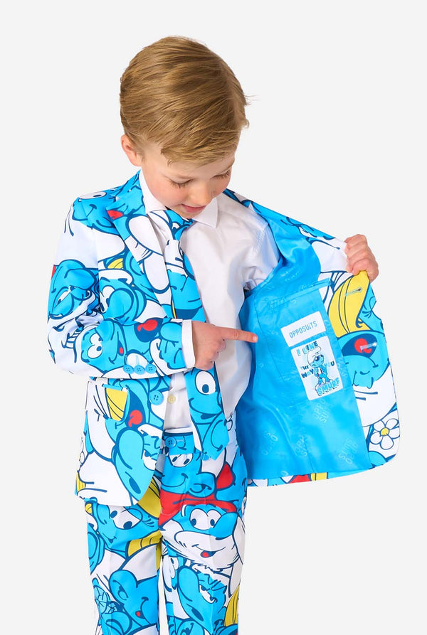 Young boy wearing a colorful Smurfs-themed kids suit featuring vibrant blue cartoon character prints, including jacket, pants, and matching tie, smiling against a plain white background. Holding one side of the jacket open.