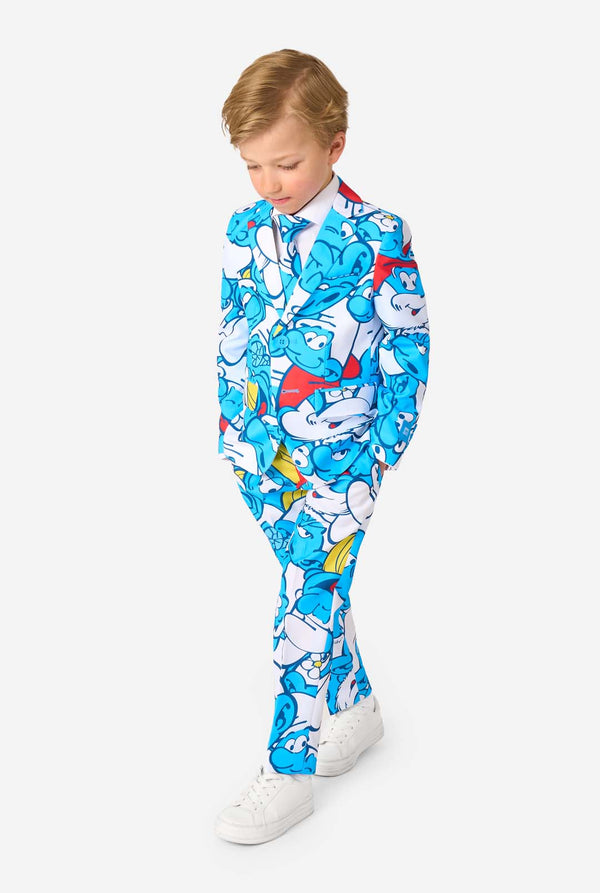 Young boy wearing a colorful Smurfs-themed kids suit featuring vibrant blue cartoon character prints, including jacket, pants, and matching tie, smiling against a plain white background. Full body, looking at the floor.