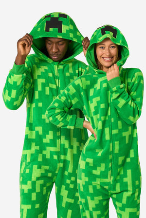 Two people wearing green pixelated onesies designed to look like the Minecraft Creeper character. The onesies have hoods with the Creeper’s iconic black pixel face. The man is holding his hood and looking down, while the women is smiling at the camera with one hand on their hip