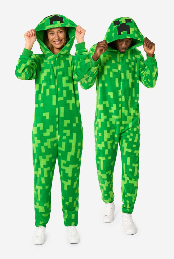 Two people wearing green pixelated onesies designed to look like the Minecraft Creeper character. The onesies have hoods with the Creeper’s iconic black pixel face. The man is holding his hood and looking down, while the woman is holding her hood and looking in the camera