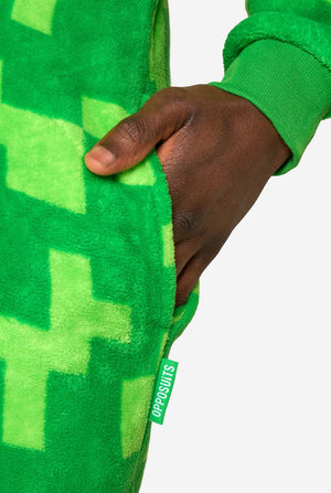 A man wearing green pixelated onesie designed to look like the Minecraft Creeper character. The onesie has a hood with the Creeper’s iconic black pixel face. The man has one hand in his pocket, close up on the pocket.