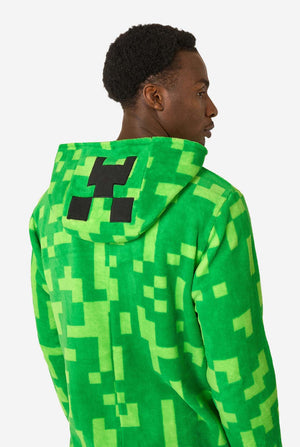 A man wearing green pixelated onesie designed to look like the Minecraft Creeper character. The onesie has a hood with the Creeper’s iconic black pixel face. The man is looking to the side. View from the back.