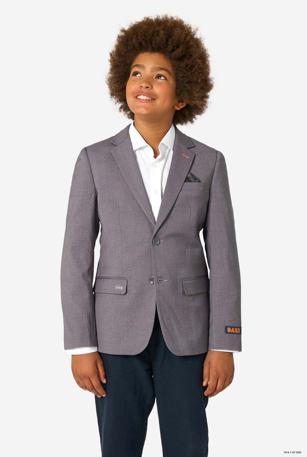 A child smiling while wearing a light grey blazer, white shirt, and dark trousers, looking upward.
