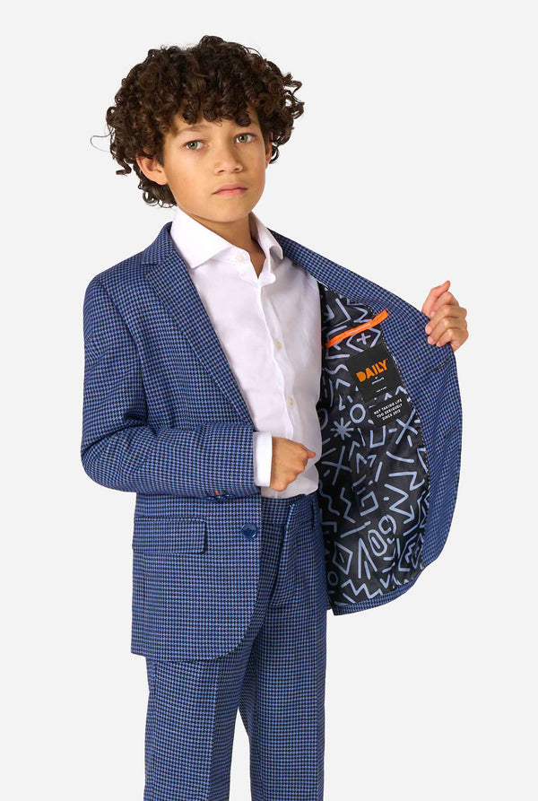 A boy wearing a navy and blue checkered suit with a white dress shirt. The suit has a modern fit with two buttons and flap pockets. He stands against a plain white background. Holding one side of the jacket open.