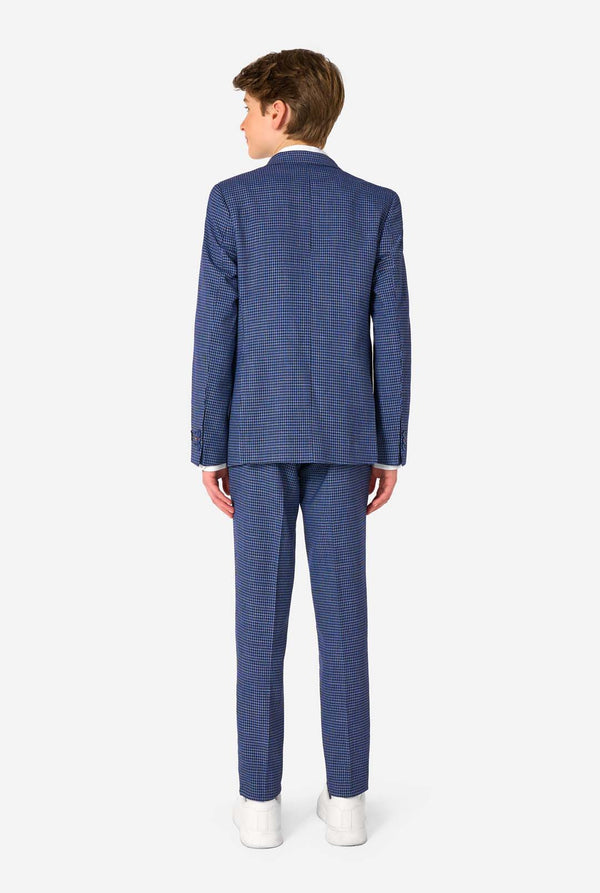 A young boy stands smiling while wearing a formal navy plaid suit jacket over a white button-down shirt. The suit features a subtle check pattern. View from the back
