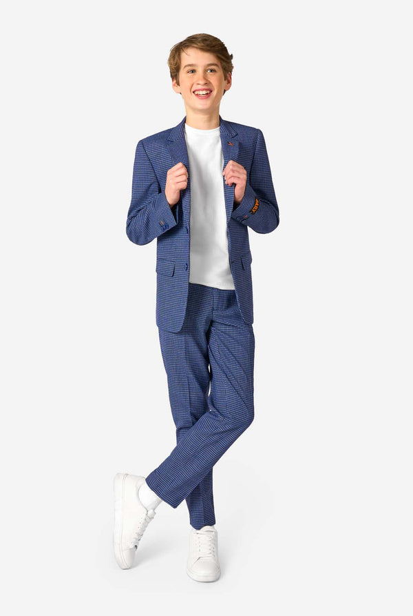 A young boy stands smiling while wearing a formal navy plaid suit jacket over a white button-down shirt. The suit features a subtle check pattern. Full body image