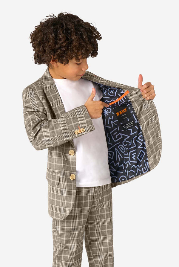 A boy wearing a tan checkered suit with a white dress shirt. The suit has a modern fit with two buttons and flap pockets. He stands against a plain white background. Holding one side of the jacket open.