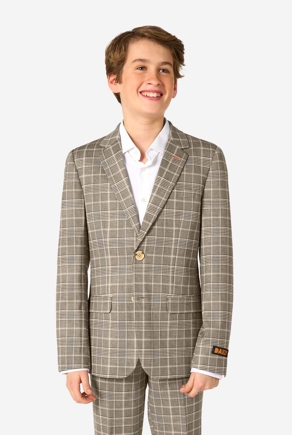 A young boy stands smiling while wearing a formal tan and beige plaid suit jacket over a white button-down shirt. The suit features a subtle check pattern.