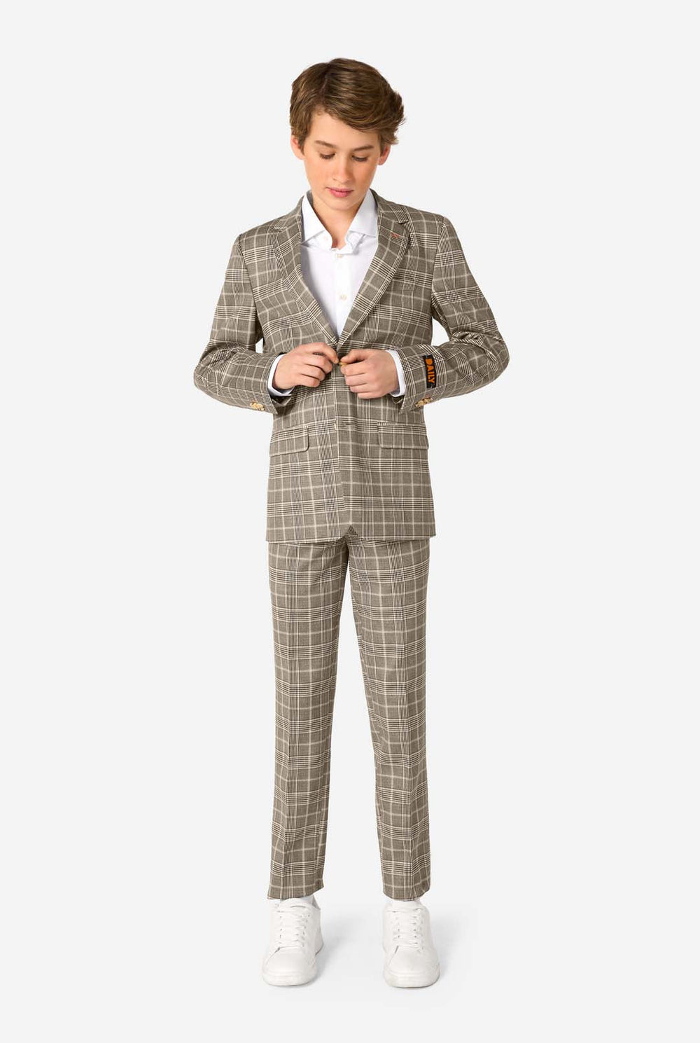 A young boy stands smiling while wearing a formal tan and beige plaid suit jacket over a white button-down shirt. The suit features a subtle check pattern.