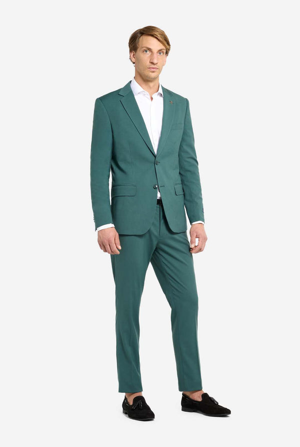 Image with light grey background and man wearing dark green men’s suit and white shirt. Suits consists of blazer and pants. Man looking towards the camera. Full body image.