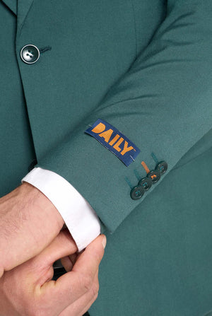Image with light grey background and man wearing dark green men’s suit and white shirt. Suits consists of blazer and pants. Sleeve close up.