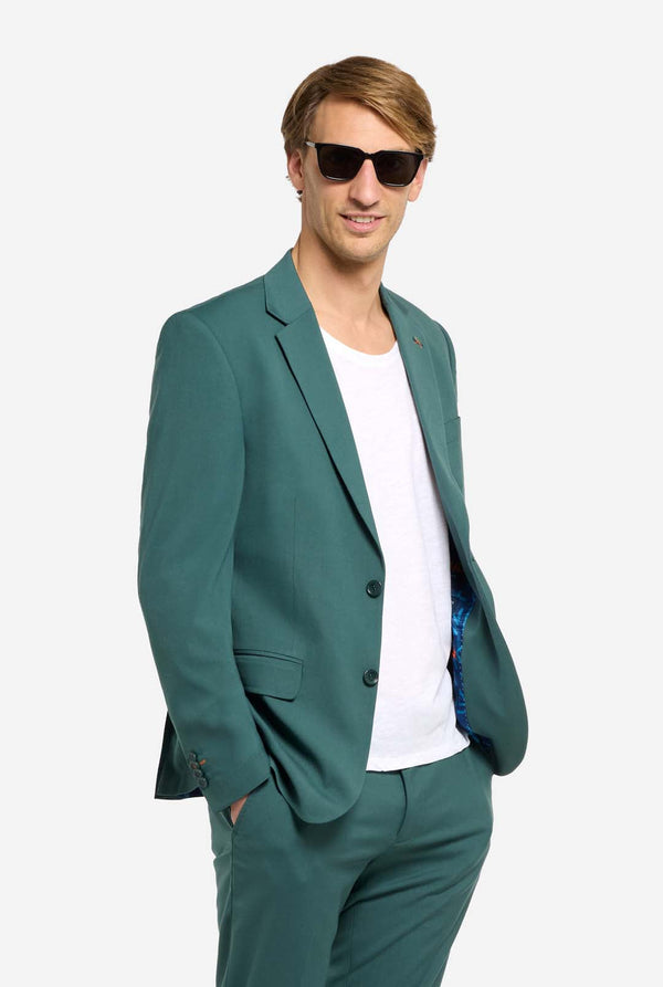 Image with light grey background and man wearing dark green men’s suit and white t-shirt. Suits consists of blazer and pants. Man looking toward the camera, and wearing sunglasses.