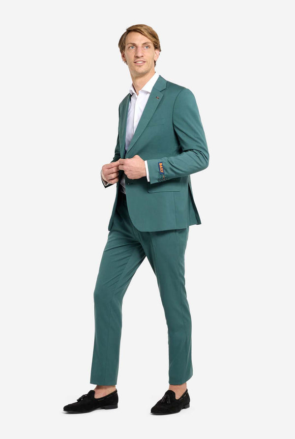 Image with light grey background and man wearing dark green men’s suit and white shirt. Suits consists of blazer and pants. Man looking side ways. Full body image.