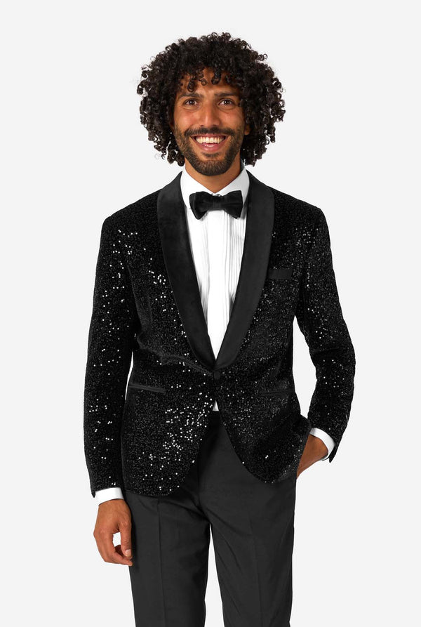 A man poses in a black sequined blazer or dinner jacket, wearing a solid white shirt underneath.