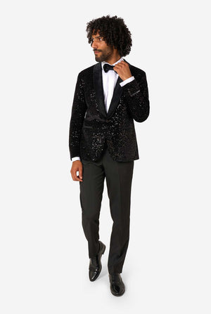 A man poses in a black sequined blazer or dinner jacket, wearing a solid white shirt underneath. Full body image