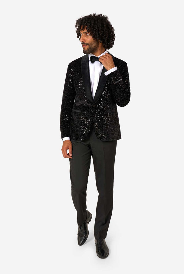 A man poses in a black sequined blazer or dinner jacket, wearing a solid white shirt underneath. Full body image