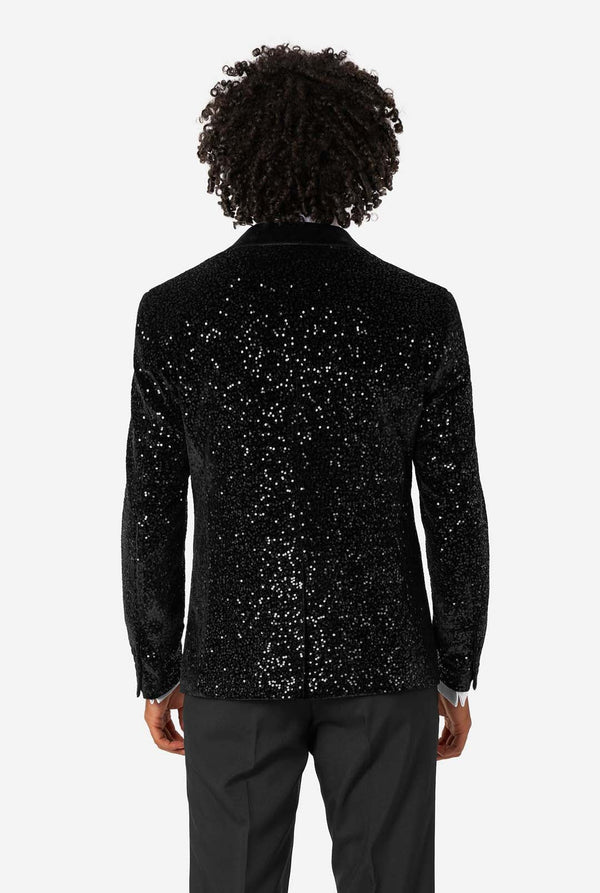 A man poses in a black sequined blazer or dinner jacket, wearing a solid white shirt underneath. View from the back
