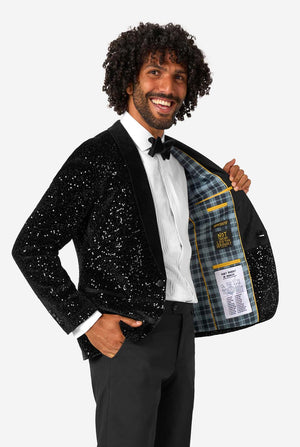A man poses in a black sequined blazer or dinner jacket, wearing a solid white shirt underneath. Showing the inside of the jacket