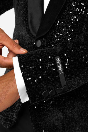 A man poses in a black sequined blazer or dinner jacket, wearing a solid white shirt underneath. Close up of the sleeves