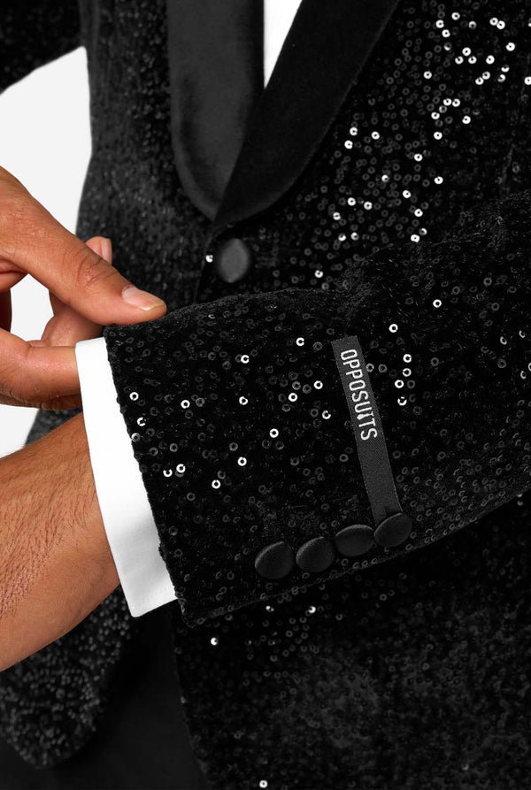 A man poses in a black sequined blazer or dinner jacket, wearing a solid white shirt underneath. Close up of the sleeves