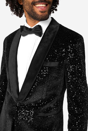 A man poses in a black sequined blazer or dinner jacket, wearing a solid white shirt underneath. Close up of the chest