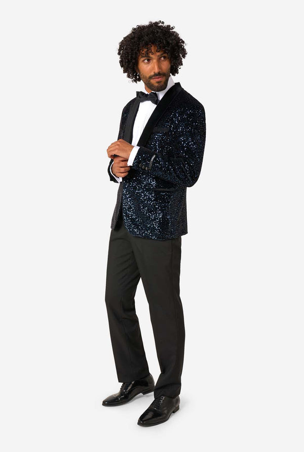 A man poses in a navy sequined blazer or dinner jacket, wearing a solid white shirt underneath. Full body image