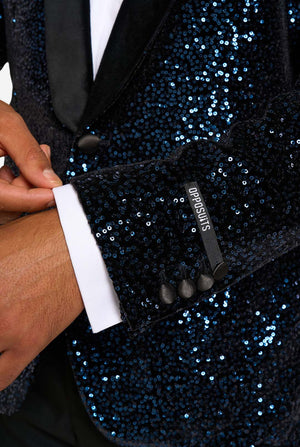 A man poses in a navy sequined blazer or dinner jacket, wearing a solid white shirt underneath. Close up of the sleeves