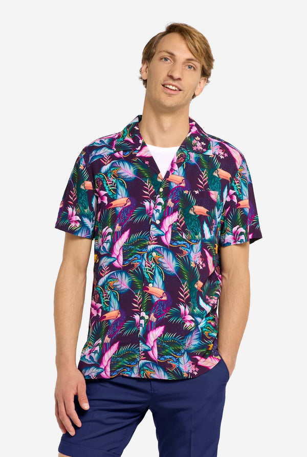 A man wearing a colorful short-sleeve Hawaiian shirt with a vibrant jungle pattern featuring tropical birds, leaves, and flowers in shades of pink, blue, green, and purple. He has light brown hair, is smiling slightly, and is standing with one hand in his pocket against a plain light background.