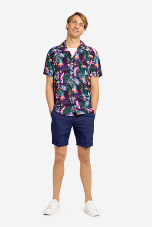 A man wearing a colorful short-sleeve Hawaiian shirt with a vibrant jungle pattern featuring tropical birds, leaves, and flowers in shades of pink, blue, green, and purple. He has light brown hair, is smiling slightly, and is standing with one hand in his pocket against a plain light background. Full body image.