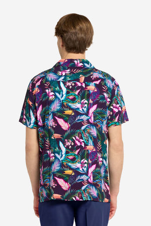 A man wearing a colorful short-sleeve Hawaiian shirt with a vibrant jungle pattern featuring tropical birds, leaves, and flowers in shades of pink, blue, green, and purple. He has light brown hair, is smiling slightly, and is standing with one hand in his pocket against a plain light background. View from the back.