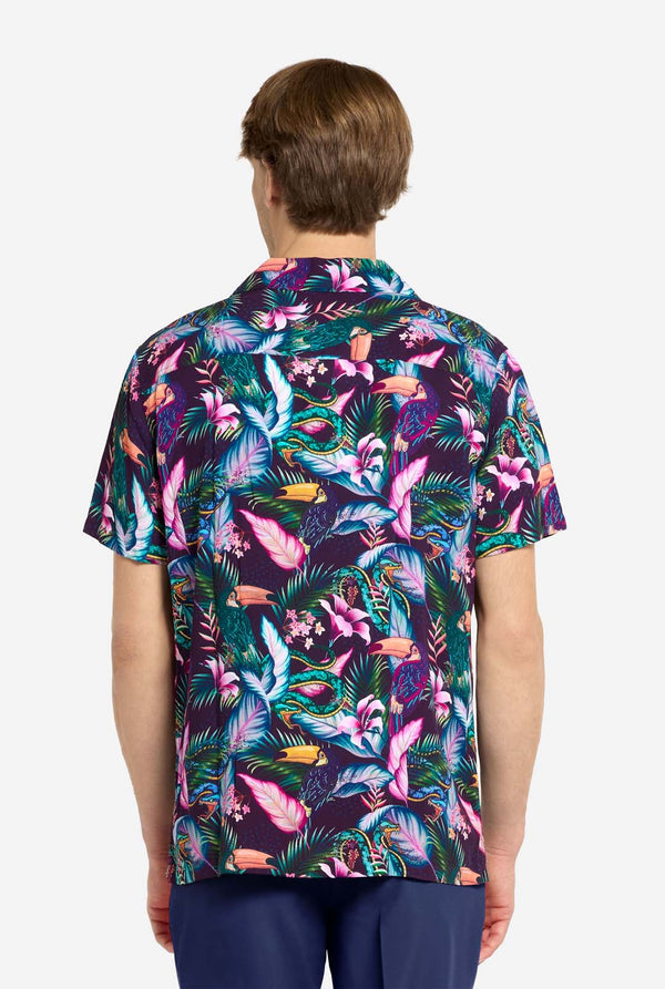 A man wearing a colorful short-sleeve Hawaiian shirt with a vibrant jungle pattern featuring tropical birds, leaves, and flowers in shades of pink, blue, green, and purple. He has light brown hair, is smiling slightly, and is standing with one hand in his pocket against a plain light background. View from the back.