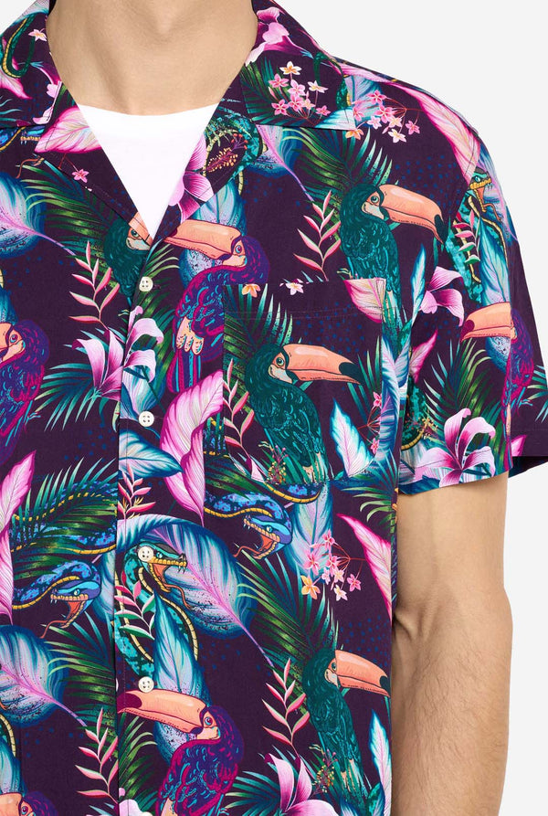 A man is wearing a purple Hawaiian shirt with a tropical print, resembling the jungle night live. Print consists of parrots, snakes, leaves and flowers. Also wearing pink shorts. He is smiling in the camera and holding one hand in the pocket. Close up.