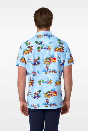 A man smiles while wearing a light blue, short-sleeved Hawaiian-style shirt printed all over with various scenes and characters from the Super Mario video game series. View from the back