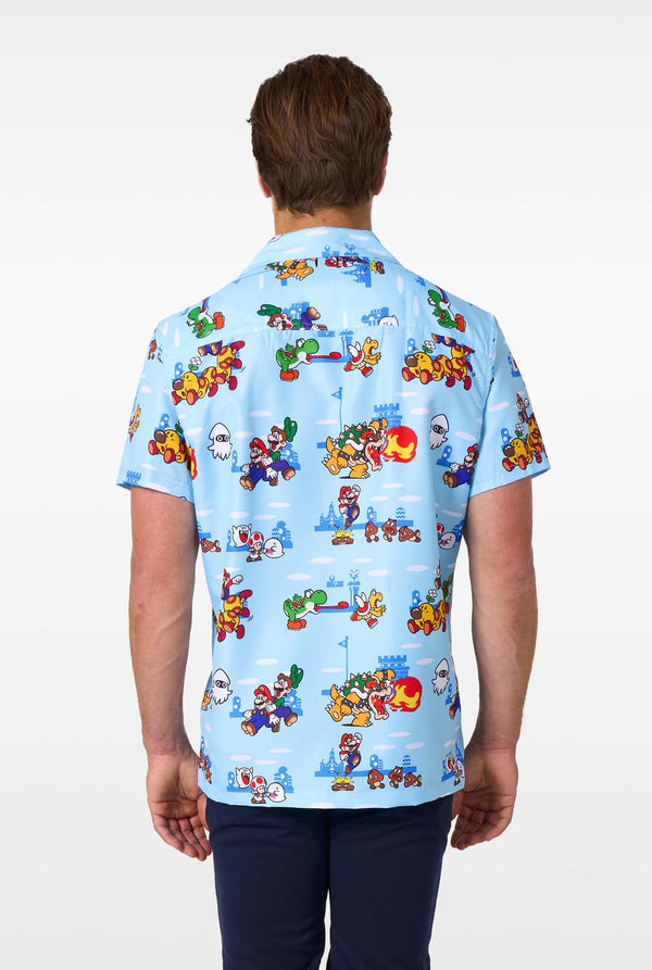 A man smiles while wearing a light blue, short-sleeved Hawaiian-style shirt printed all over with various scenes and characters from the Super Mario video game series. View from the back