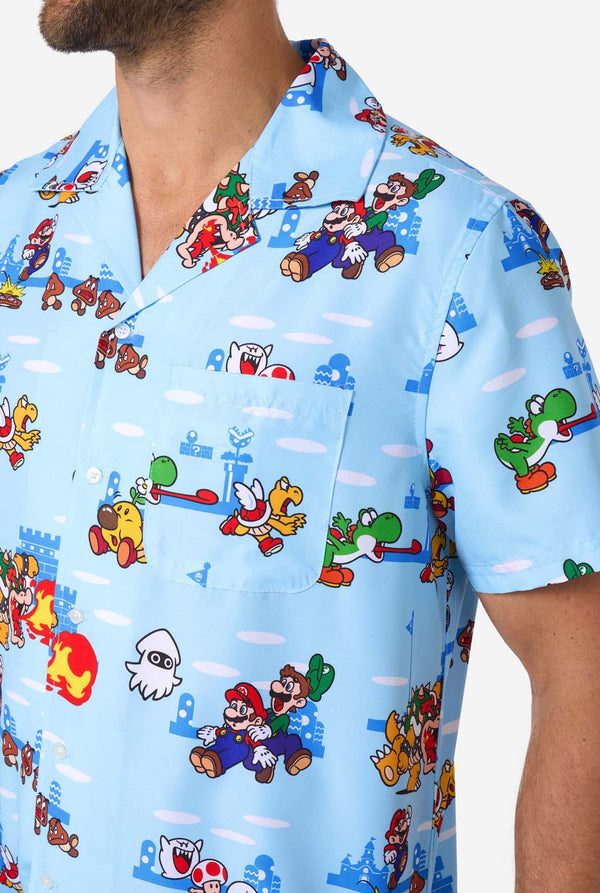 A man smiles while wearing a light blue, short-sleeved Hawaiian-style shirt printed all over with various scenes and characters from the Super Mario video game series. Close up of the chest