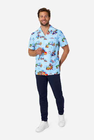 A man smiles while wearing a light blue, short-sleeved Hawaiian-style shirt printed all over with various scenes and characters from the Super Mario video game series. Full body image