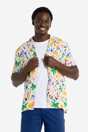 A man is wearing a white Hawaiian shirt with a rainbow pride print. The Print consists of peace symbols, hearts, and other icons. He is also wearing blue pants. He is smiling at the camera and holding the sides of the shirts.