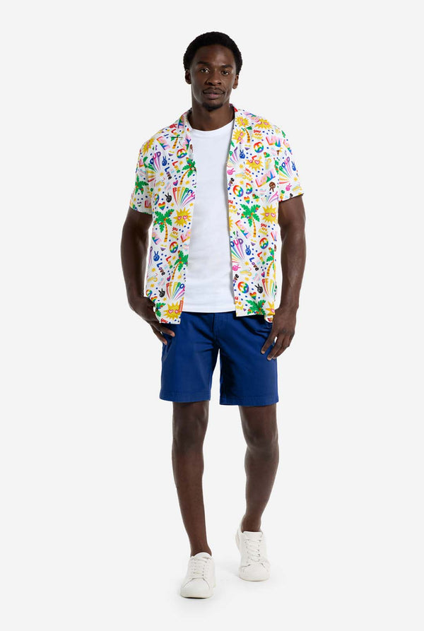 A man is wearing a white Hawaiian shirt with a rainbow pride print. The Print consists of peace symbols, hearts, and other icons. He is also wearing blue pants. He is smiling at the camera and holding the sides of the shirt. Full body image.