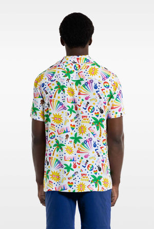 A man is wearing a white Hawaiian shirt with a rainbow pride print. The Print consists of peace symbols, hearts, and other icons. He is also wearing blue pants. He is smiling at the camera and holding the sides of the shirt. View from the back.